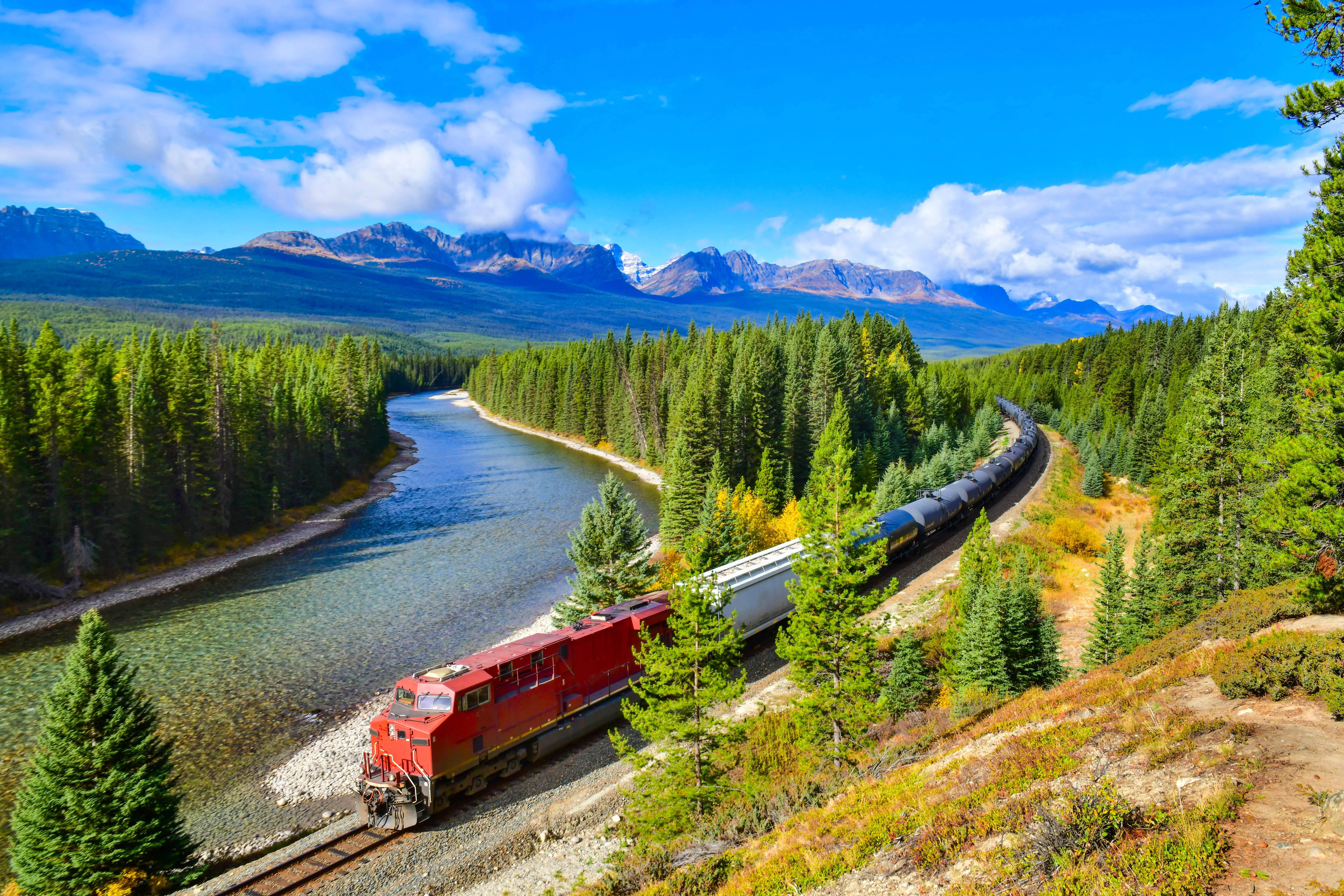 Train History: A red locomotive travelling along the Canadian Pacific Railway, winding alongside a scenic river with dense evergreen forests and towering mountains in the background