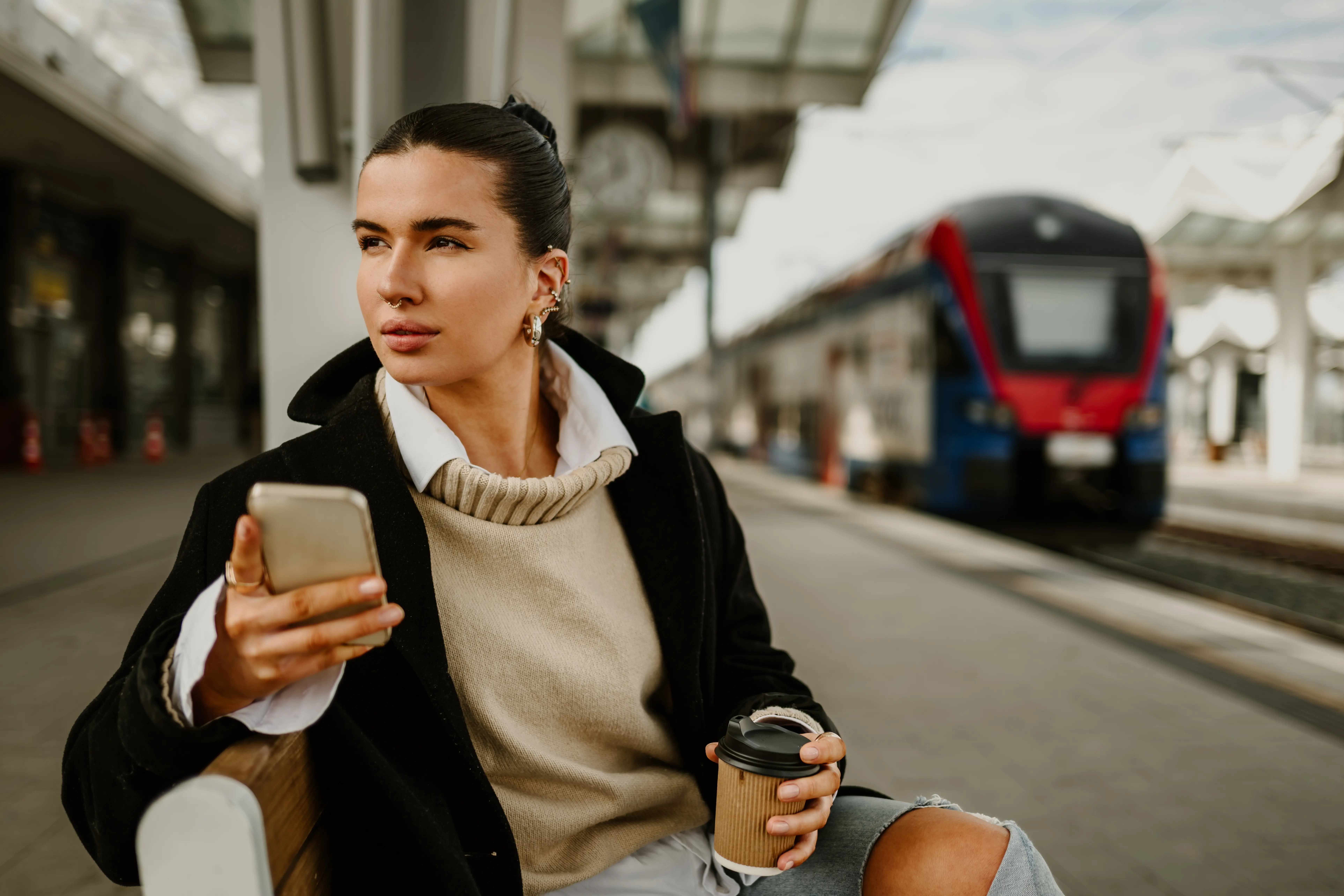 Train Timetables: A female commuter sitting down at a train station with her phone in one hand and a coffee in the other.